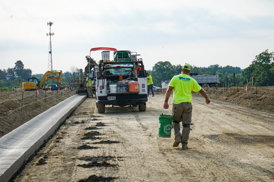 Construction worker walking along a subdivision road beside newly poured concrete curb, with a work truck, excavator, and other construction equipment in the background.